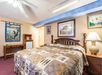 A bedroom with a large bed, patterned quilt, nightstand with lamp, wall art, wooden desk, and mini-fridge. Ceiling fan and light overhead.