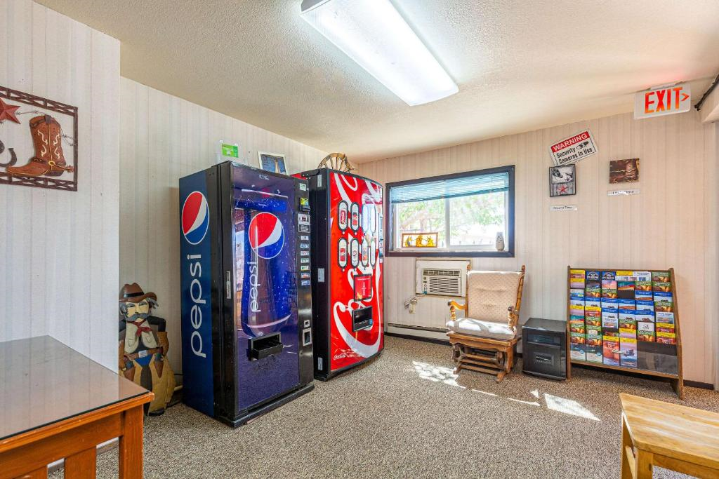 Room with a Pepsi vending machine, a Coke machine, brochures on a stand, and a wooden chair. An air conditioner is under a window. Cowboy decor on the wall. Exit sign above door.