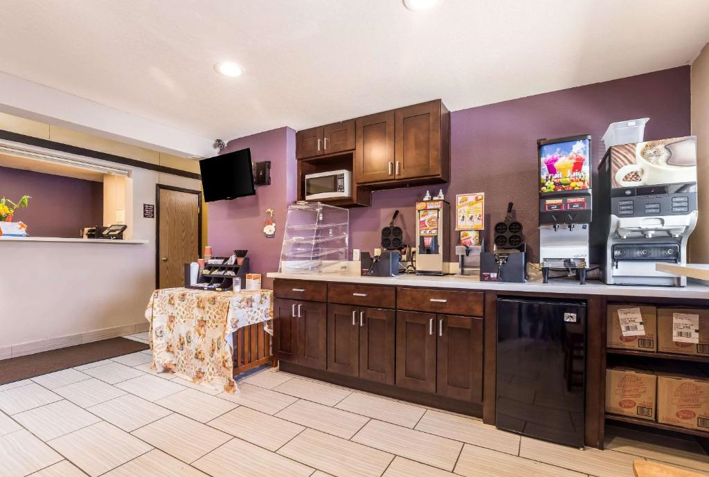 Breakfast area with wooden cabinets, waffle makers, beverage dispensers, and a TV mounted on the wall. Counter with a floral cloth holds food items. Tile flooring and purple walls in the background.