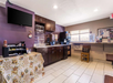 Hotel breakfast area with a counter and cabinets, a mounted TV, a table with a floral tablecloth, beverage dispensers, and a wall-mounted brochure display near a window.
