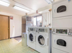 Laundry room with three stacked washers and dryers. Fluorescent lighting, a window, and a wooden door are visible. Floors have a pale green tile pattern.