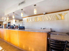 Hotel lobby with a front desk featuring a "Rodeway" sign on the wall. The area is well-lit with hanging lights and has a granite countertop with a few electronic devices.