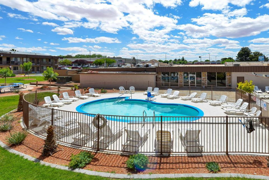 Outdoor pool area with loungers and tables, surrounded by a metal fence. Building and parking lot visible in background, under a partly cloudy sky.