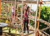 Two people wearing harnesses navigate ropes and obstacles on an outdoor ropes course structure, surrounded by trees.