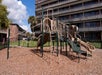 Playground equipment with slides and climbing structures on woodchip ground, set against a multi-story building.