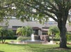 A fountain in front of a beige building with lush greenery and palm trees surrounding it. A sign is visible on the building.