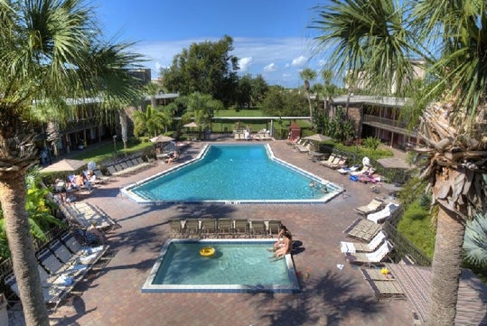 Aerial view of a hotel courtyard with a large, central pool surrounded by lounge chairs and palm trees. A smaller, circular pool is in the foreground. Several people are relaxing by the pools.