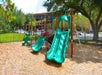 Playground with two green slides, climbing structures, and a swing set on a bed of woodchips, surrounded by trees and a metal fence.