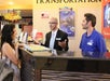 A woman stands at a hotel reception desk speaking with two male employees. One employee gestures with his hands while the other listens. Various promotional signs are visible in the background.