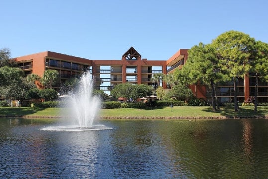 A modern hotel building stands behind a pond with a central fountain, surrounded by trees and landscaped gardens under a clear blue sky.