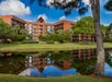 A large, multi-story brick building with balconies is reflected in a pond. Tall trees and green grass surround the area under a blue sky with clouds.