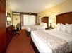 Hotel room with two double beds, a desk with a chair, and a TV on a cabinet. A window with patterned curtains overlooks greenery. Warm lighting and neutral-toned decor are present.