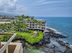 Aerial view of a coastal hotel surrounded by palm trees and situated on a rocky shoreline, with the ocean extending into the distance under a cloudy sky.