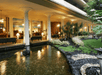Lobby area with columns and seating, overlooking a pond with stones and plants, surrounded by greenery. Warm lighting illuminates the space.