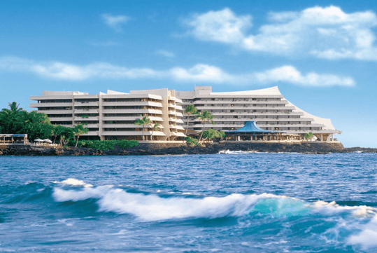 Large waterfront hotel with angular architecture, surrounded by palm trees, against a backdrop of blue sky and ocean waves.