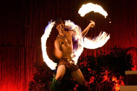Person performing a fire dance at night, with two circles of fire swirling around them, wearing a traditional costume against a backdrop of bamboo and foliage.