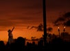 A woman in traditional attire performs with her arm raised against a sunset backdrop, with silhouetted trees and tiki torches in the foreground.