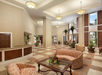 Hotel lobby with high ceilings, beige walls, checkered floor, and modern lighting. Features patterned seating, glass coffee table with flowers, and large windows with plants.