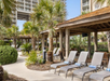 Poolside lounge chairs with striped towels under wooden cabanas, surrounded by palm trees and greenery, near a tall building.