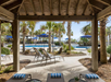 View from a shaded cabana of an outdoor pool area with palm trees, lounge chairs, and blue umbrellas on a sunny day.