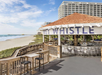 Outdoor beachside bar with "Whistle" sign, wooden deck, and seating. Located near a sandy shoreline with a high-rise building in the background under a clear blue sky.