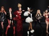 Six drag performers stand in a row, each wearing distinct, elaborate costumes in various styles and colors, posing confidently against a dark background.