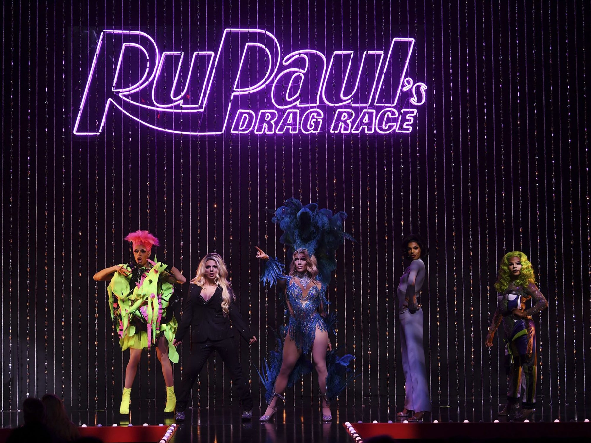 Five drag queens pose on stage in colorful costumes beneath a large illuminated "RuPaul's Drag Race" sign, with vertical light strands in the background.