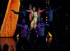 A woman in a yellow and green bodysuit performs on stage with five shirtless male dancers, all striking dynamic poses under stage lighting.