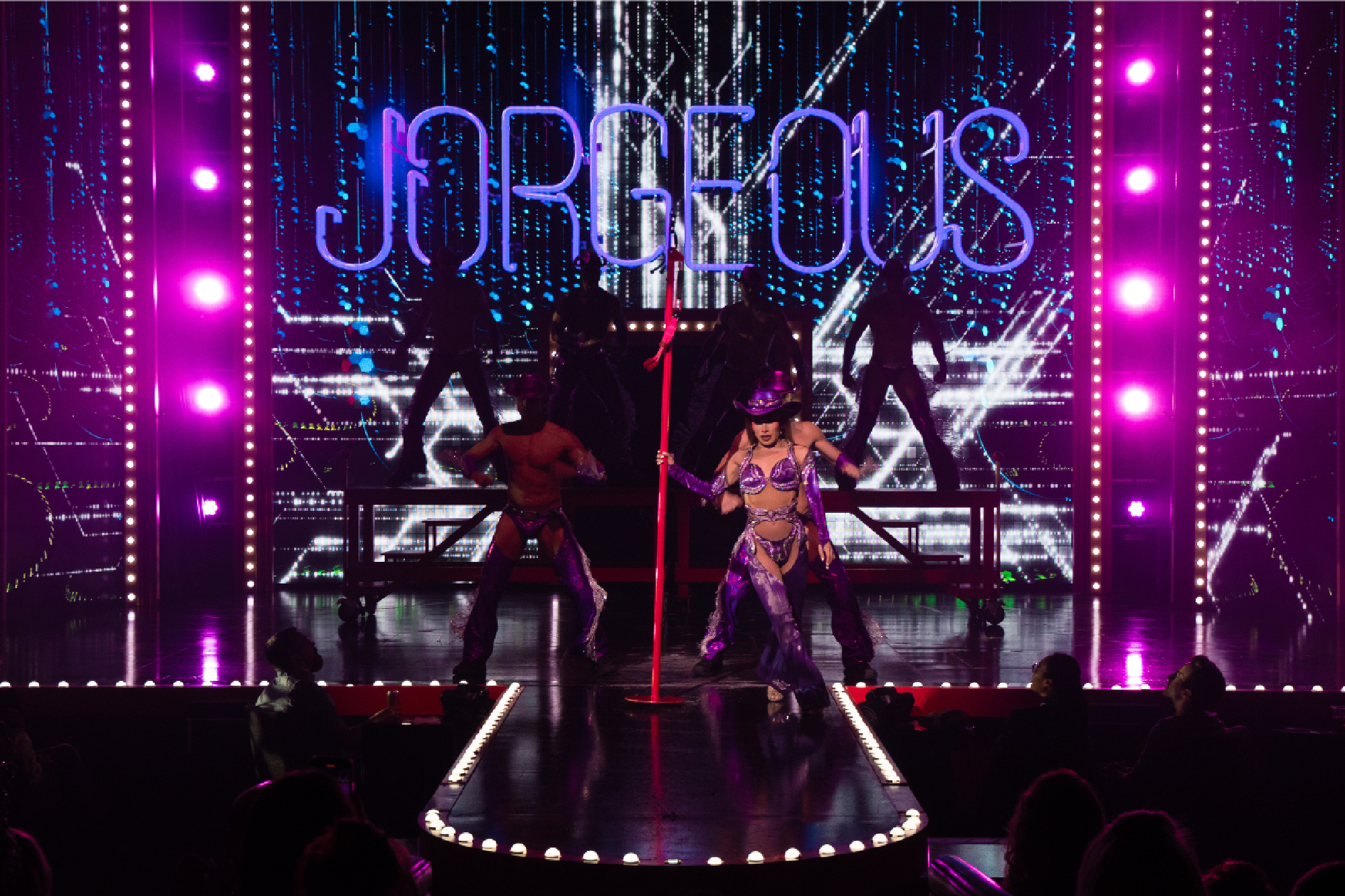 Performer in a purple costume dances on stage with a pole, backed by dancers in silhouette. Neon "GORGEOUS" sign and pink-purple lights illuminate the scene.