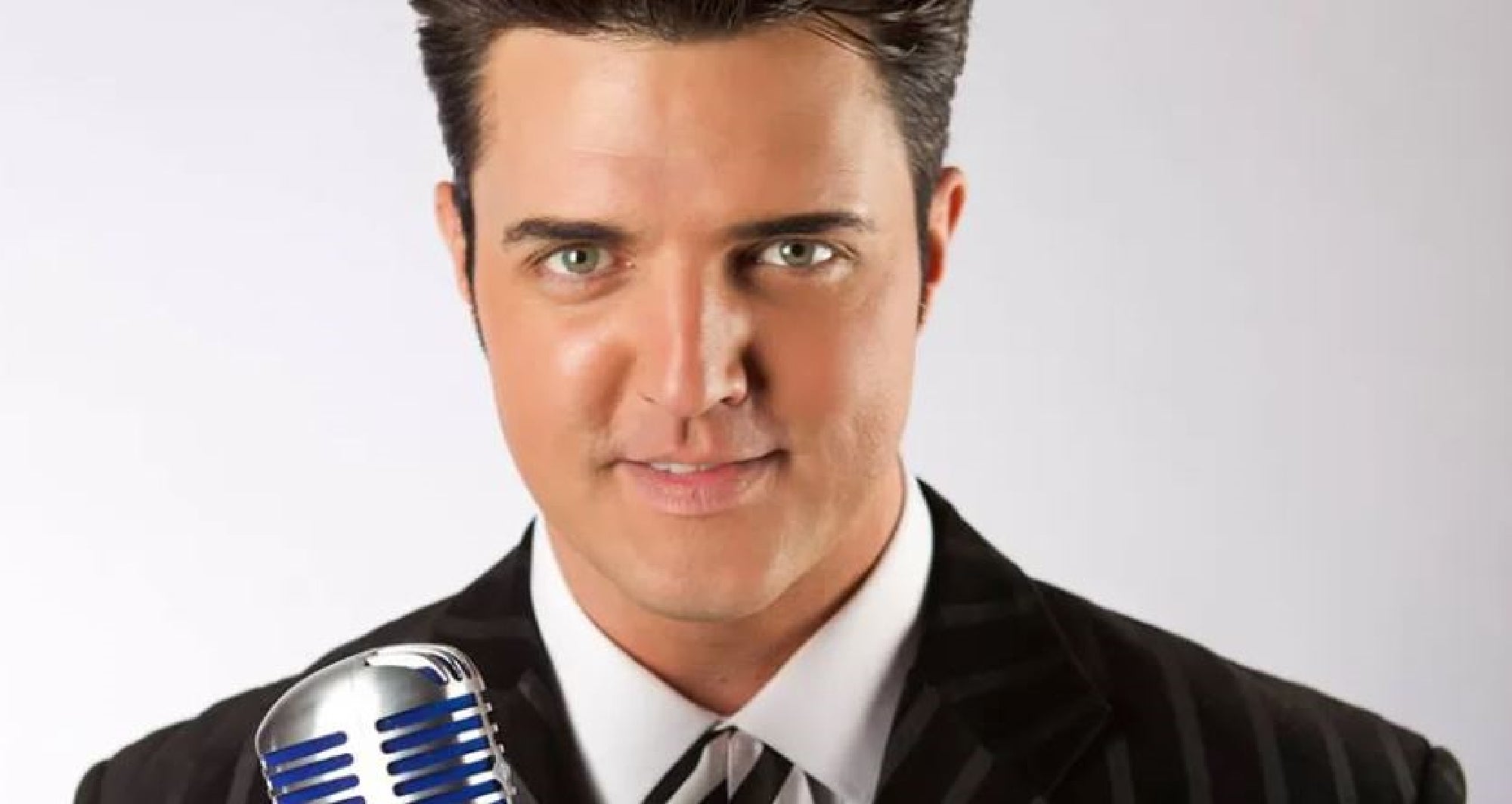 Man in a dark striped suit holds a vintage microphone, smiling at the camera against a plain background.