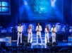 Five performers in white outfits sing on stage with a blue nautical backdrop. The scene includes musicians in the background and stage lights illuminating the performers.