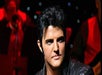 A person in a leather jacket is on stage, looking to the side, with a blurred background of musicians and lights.