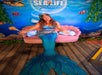 A person dressed as a mermaid sits on a pink shell-shaped couch in front of a Sea Life Orlando Aquarium backdrop.