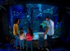 A family, including a woman and four children, watching divers interact with colorful fish and a sea turtle in an aquarium, while one of the adults points out something of interest.