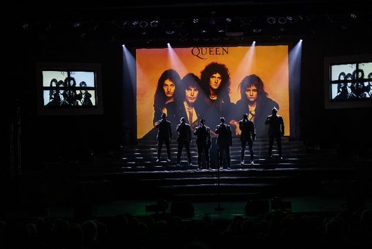 The Six Brothers performing on stage with an image of the iconic rock band Queen displayed on the LED screen as their backdrop.