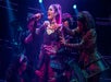 Performer in a studded costume sings passionately on stage, surrounded by dancers in matching outfits.