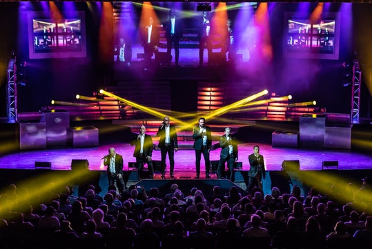 Five performers in suits sing on a brightly lit stage with colorful lights and smoke effects, while an audience watches. Two large screens show a close-up of the performers.