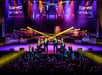 Five performers in suits sing on a brightly lit stage with colorful lights and smoke effects, while an audience watches. Two large screens show a close-up of the performers.