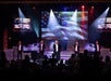 Five men perform on stage with microphones; large American flags are displayed on screens behind them. There is stage lighting and an audience watching the performance.