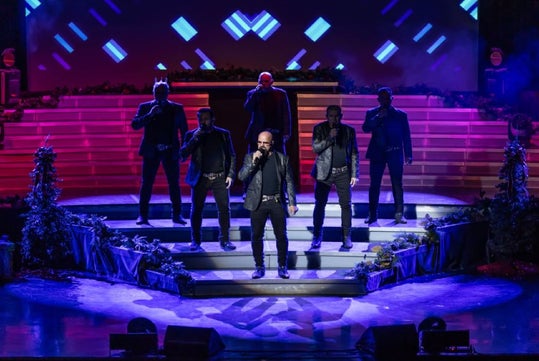 Five men stand on a multi-level stage singing into microphones under blue and purple lighting, with geometric light patterns in the background.