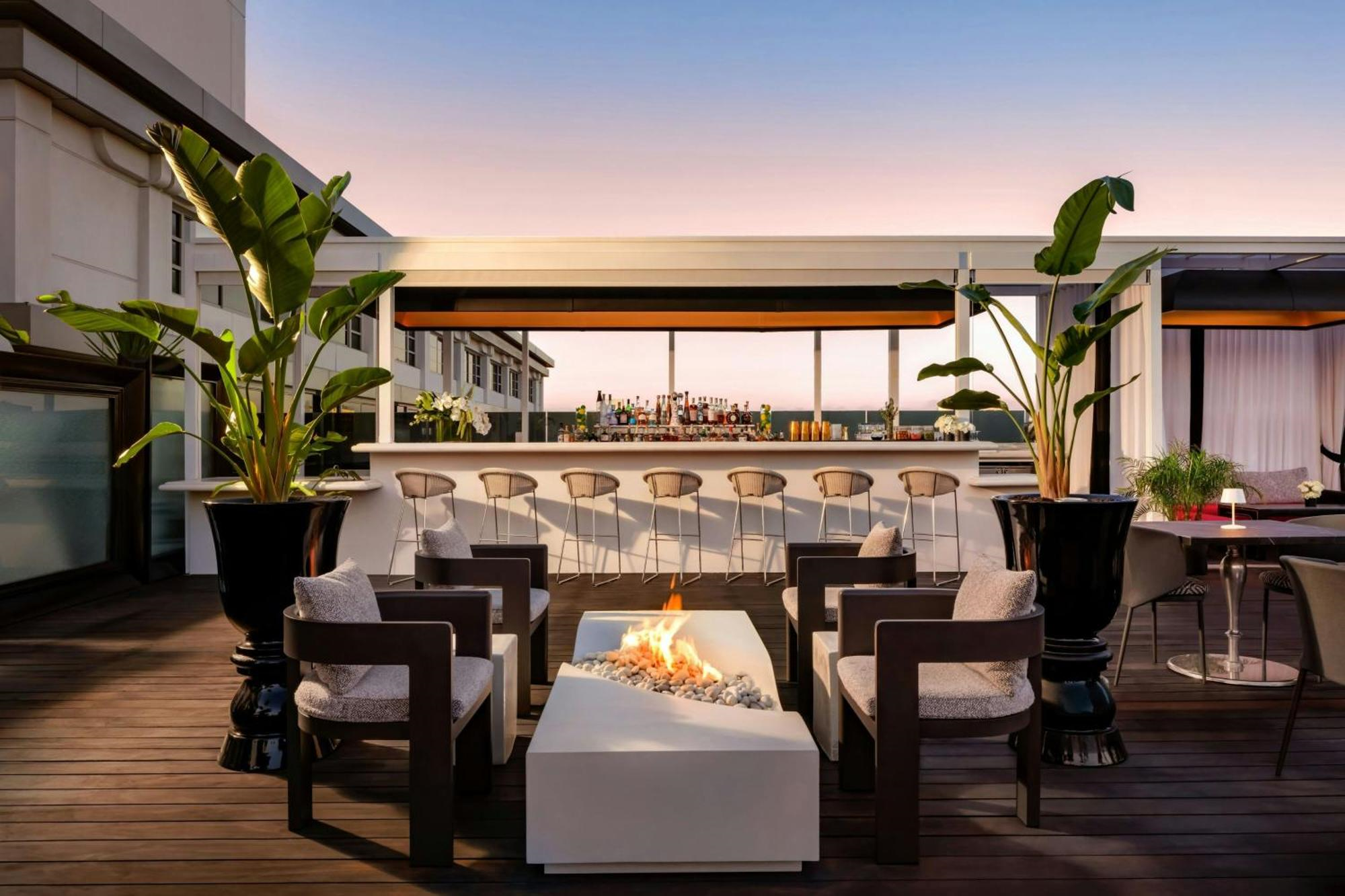 A modern rooftop lounge with chairs around a central firepit, tall potted plants, and a bar with stools in the background at sunset.