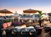 Modern rooftop lounge with fire pit, cushioned seating, round stools, potted plants, and outdoor dining tables under large umbrellas at sunset.