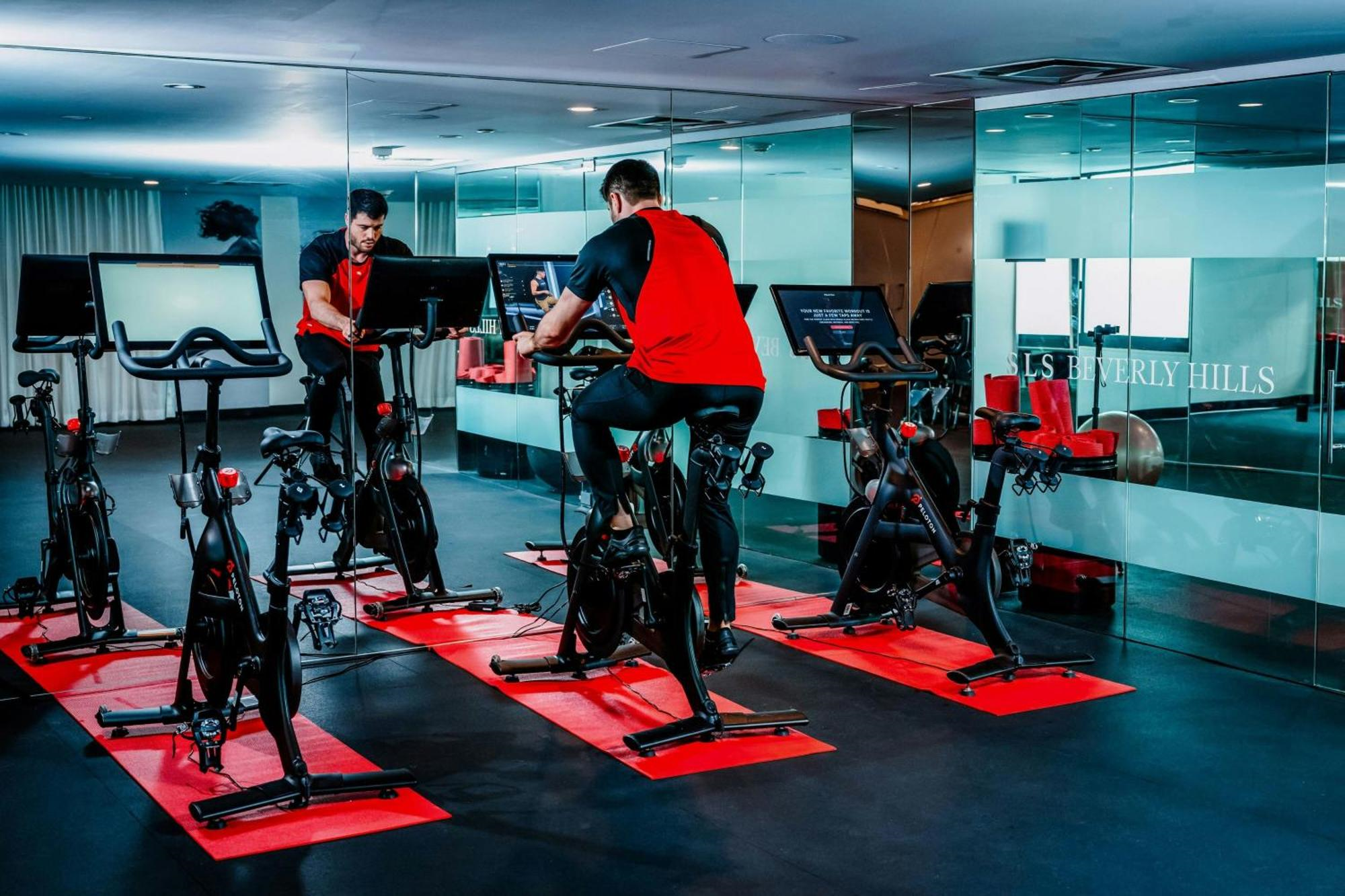 Two people in red shirts use stationary bikes with screens in a modern gym with mirrors and several cycling stations on red mats.