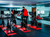 Two people in red shirts use stationary bikes with screens in a modern gym with mirrors and several cycling stations on red mats.