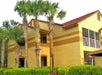 Two-story yellow building with terracotta roof and arched windows, surrounded by palm trees and green bushes.