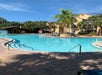 Outdoor pool area with clear water, surrounded by palm trees and a few lounge chairs. Multi-story beige building visible in the background under a clear blue sky.