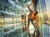A child in a brown jacket and green pants poses on a mirrored floor in a glass room. Two people in the background are walking.