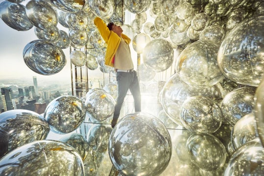 Person standing inside a room filled with reflective silver spheres, stretching with arms behind head. Sunlight streams through large windows, illuminating the scene. Cityscape visible outside.