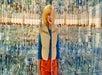 Person with curly hair stands in a reflective, mirrored room, wearing a beige vest, blue sweater, and orange pants. The environment creates multiple reflections of the person and surroundings.