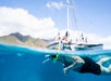 A person snorkeling near a catamaran boat with several people onboard; mountains and blue sky are visible in the background.